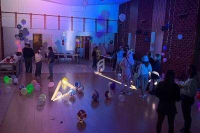 Salle décorée avec des ballons colorés, éclairée par des lumières tamisées, avec des personnes discutant et se déplaçant.