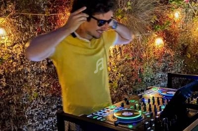 Un DJ en chemise jaune mixe de la musique sur une console éclairée, entouré de lumières et de végétation.