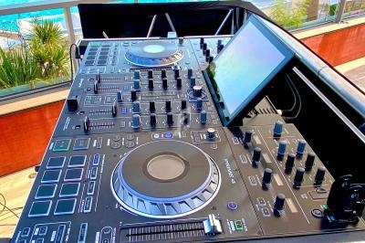 Table de mixage DJ installée en extérieur avec vue sur une piscine, des transats et des arbres en arrière-plan.