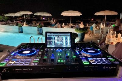 Table de mixage DJ éclairée près d'une piscine, avec des invités assis sous des parasols en arrière-plan.