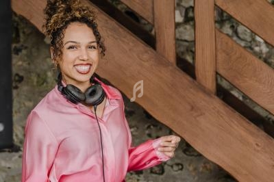 Une femme souriante en chemisier rose mixe de la musique sur une table de mixage, devant un mur en pierre.
