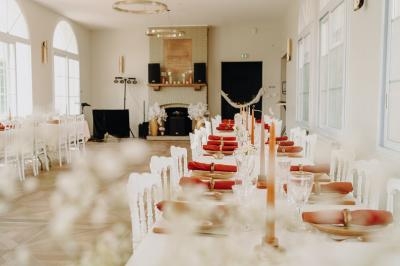 Salle de réception lumineuse avec tables décorées de nappes blanches, bougies et serviettes rouges.