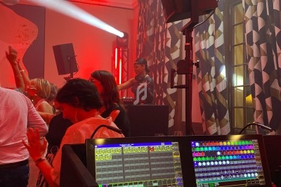 Des personnes dansent devant un DJ, avec une console de mixage lumineuse au premier plan et des éclairages rouges.