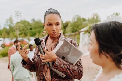 Une femme tenant un micro et une tablette discute avec une autre personne en extérieur, dans un cadre naturel.
