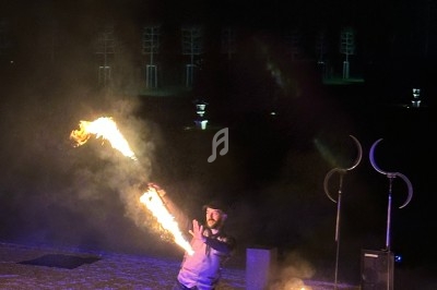 Un artiste jongle avec des bâtons enflammés dans un espace extérieur sombre, éclairé par des flammes et des lumières.