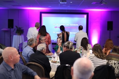 Des personnes assises et debout lors d'un événement, face à un écran projetant un message sur fond violet.