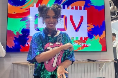 Une DJ souriante mixe sur une table de mixage devant un écran coloré dans un environnement d'exposition.