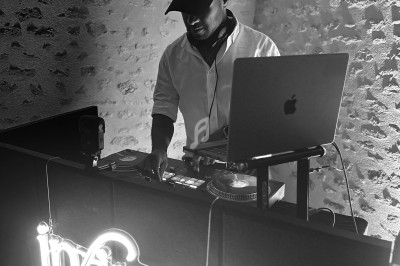 Un DJ mixe de la musique sur une table de mixage, devant un panneau lumineux ’infinia’, dans un espace en pierre.