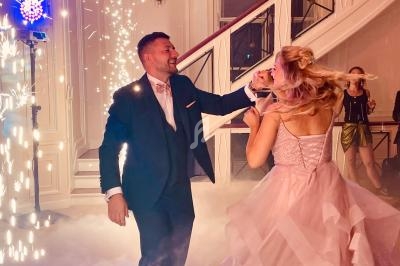 Un couple danse au milieu de la fumée et des étincelles dans une salle élégante avec un escalier en arrière-plan.