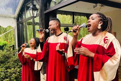 Trois chanteurs en tenue de chorale rouge et beige interprètent une chanson avec des micros en extérieur.