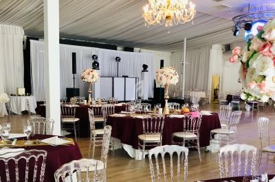 Salle de réception décorée avec des tables nappées de bordeaux, des chaises transparentes et des centres de table fleuris.