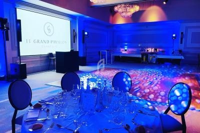 Salle de réception élégamment éclairée en bleu, avec tables dressées et écran projetant ’Le Grand Pavillon Chantilly’.
