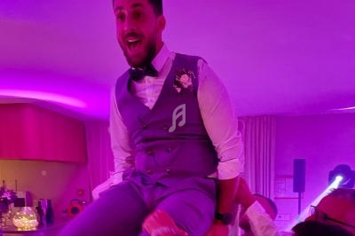 Un homme en costume violet est porté sur les épaules de quelqu'un lors d'une fête dans une salle éclairée en rose.