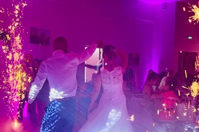 Un couple danse sous des étincelles dans une salle éclairée par des lumières violettes, entouré d'invités.