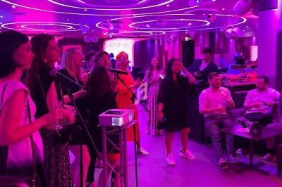 Des personnes chantent au karaoké dans une salle éclairée par des lumières roses et violettes, avec un public assis.