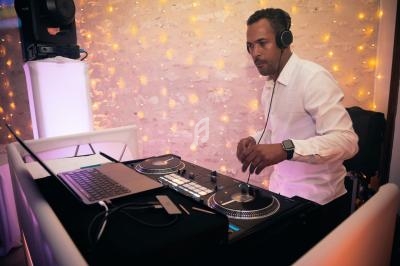 Un DJ en chemise blanche mixe de la musique sur une table de mixage, avec un mur éclairé en arrière-plan.