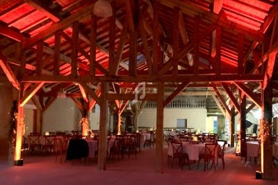 Salle en bois avec charpente apparente, tables rondes dressées et éclairage chaleureux rouge et orange.