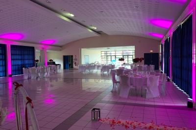 Salle de réception décorée avec des nappes blanches, éclairage rose et tables dressées pour un événement.