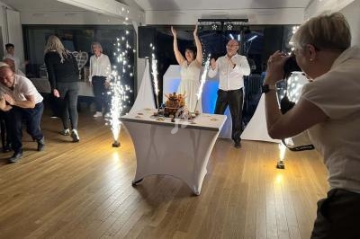 Une femme et un homme célèbrent devant un gâteau avec des étincelles, entourés de personnes dans une salle éclairée.