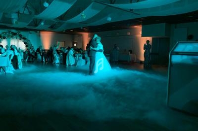 Un couple danse au centre d'une salle de réception illuminée en bleu, entouré de fumée et d'invités assis.