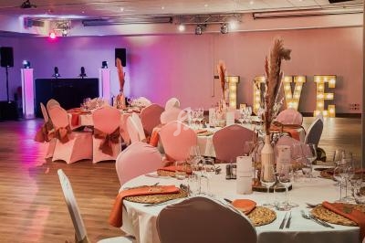 Salle de réception décorée avec des tables dressées, chaises roses et lettres lumineuses formant ’LOVE’ en arrière-plan.