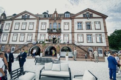 Façade d'un grand bâtiment historique en pierre avec des escaliers, des fenêtres à volets et une terrasse aménagée.