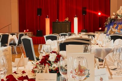 Salle de réception décorée pour un mariage, avec tables dressées, fleurs rouges et blanches, et une scène en arrière-plan.