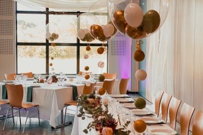 Salle décorée pour un événement avec tables dressées, ballons suspendus et guirlandes végétales.