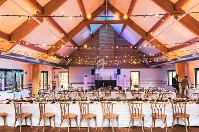 Salle de réception décorée avec des guirlandes lumineuses, tables dressées et chaises en bois sous un plafond en poutres…
