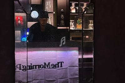 Un homme portant un chapeau se tient derrière un bar éclairé, reflété dans un miroir fragmenté.