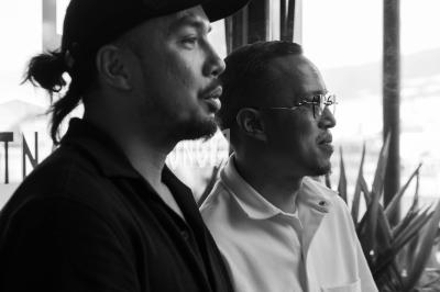 Deux hommes debout, l'un portant une casquette et l'autre des lunettes, regardant dans la même direction.