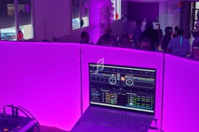 Ordinateur portable affichant un logiciel de DJ sur une table éclairée en violet, avec des invités en arrière-plan.