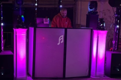 Un DJ en sweat rouge mixe derrière une table éclairée par des lumières roses dans un environnement sombre.