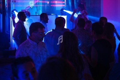 Personnes dansant sous des lumières colorées dans une ambiance festive avec un DJ en arrière-plan.