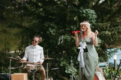 Une chanteuse en robe verte et un batteur jouent de la musique en plein air devant des arbres.