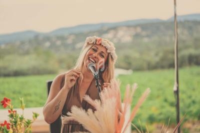 Femme chantant au micro en extérieur, portant une couronne de fleurs, avec un paysage naturel en arrière-plan.