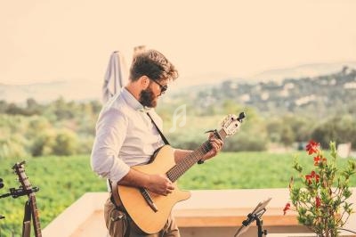 Un homme joue de la guitare acoustique en extérieur, devant un paysage verdoyant et ensoleillé.