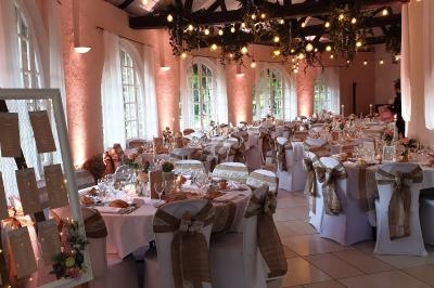 Salle de réception décorée avec des tables dressées, des chaises ornées de rubans et des lumières suspendues.