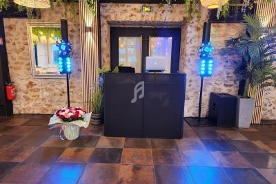 Pupitre de DJ avec ordinateur portable, éclairages colorés et décoration végétale dans une salle au mur en pierre.