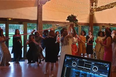 Des invités dansent et tiennent des rubans colorés autour d'une mariée sous des guirlandes lumineuses dans une salle festive.