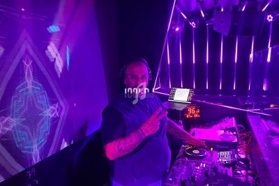 Un DJ mixe sur une table de mixage dans une ambiance lumineuse violette, faisant un signe de la main vers l'objectif.