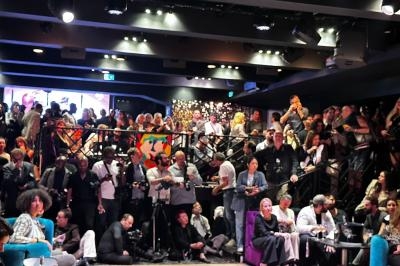 Salle bondée avec des personnes debout et assises, éclairage tamisé, ambiance animée lors d'un événement.