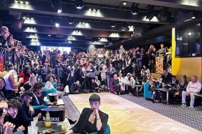 Salle bondée avec des spectateurs assis et debout autour d'une scène centrale, éclairée par des projecteurs.