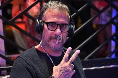 Homme portant des lunettes et un casque audio, faisant un signe de la main devant une grille métallique.