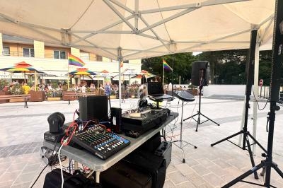 Équipement de sonorisation installé sous un chapiteau, avec une place animée et des drapeaux arc-en-ciel en arrière-plan.