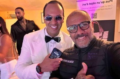 Deux hommes souriants posent pour une photo dans un environnement intérieur, avec d'autres personnes en arrière-plan.