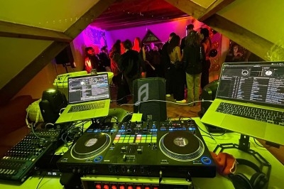 Vue d'une table de mixage avec ordinateurs et platines, face à une salle où des personnes dansent sous des lumières colorées.