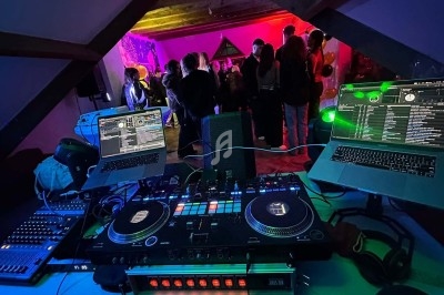 Vue d'une table de mixage avec deux ordinateurs portables, face à une fête dans une pièce éclairée par des lumières colorées.