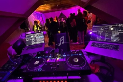 Vue d'une table de mixage avec deux ordinateurs, des platines et une fête en arrière-plan sous un éclairage coloré.