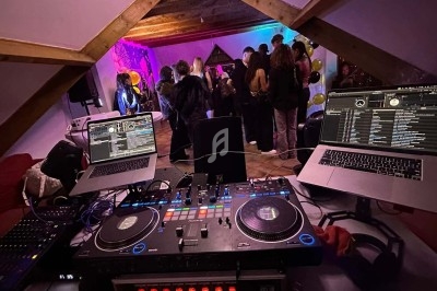 Vue depuis une table de mixage avec deux ordinateurs, face à un groupe de personnes dansant dans une pièce éclairée.